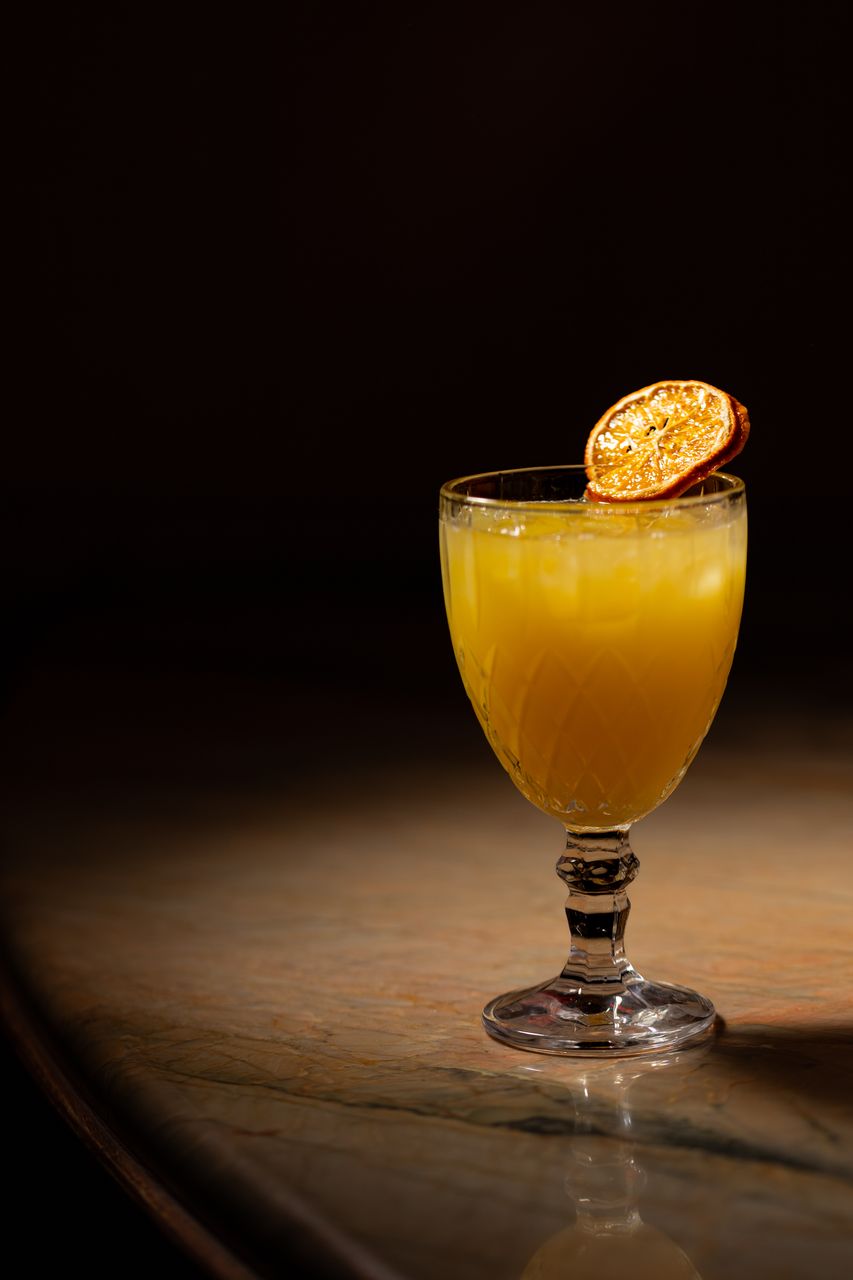 A golden-orange cocktail in a cut-crystal goblet with a dried citrus garnish